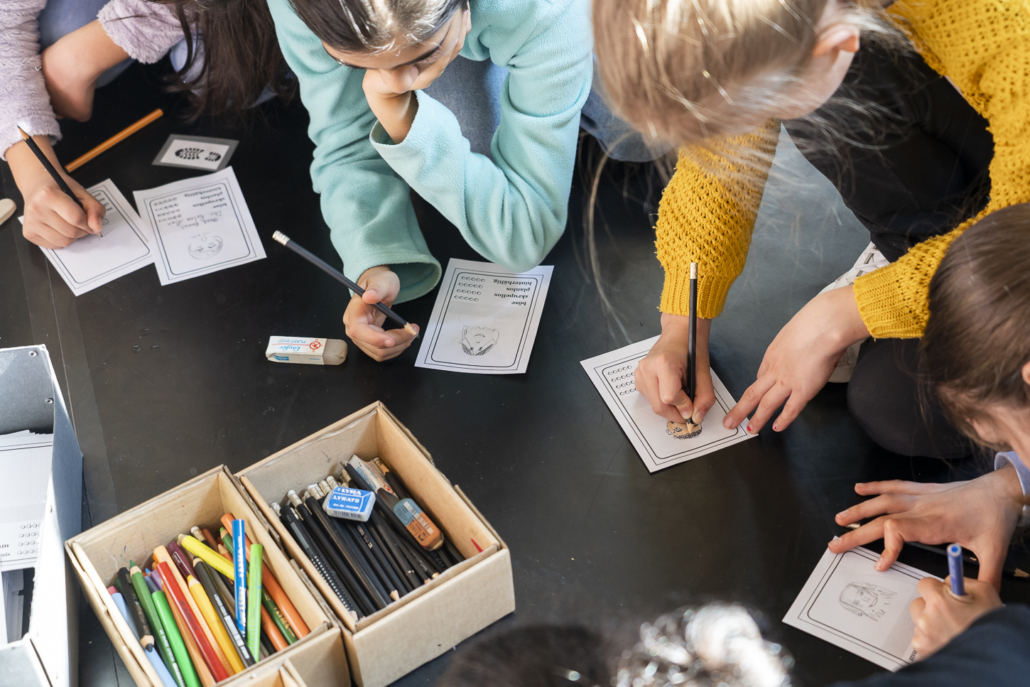 Kinder knien auf dem Boden und arbeiten im Rahmen einer Krimi-Werkstatt an ihren Schreibprojekten. Daneben stehen Boxen mit Stiften.