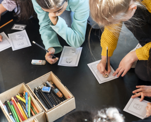 Kinder knien auf dem Boden und arbeiten im Rahmen einer Krimi-Werkstatt an ihren Schreibprojekten. Daneben stehen Boxen mit Stiften.