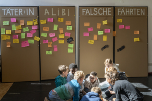 081_GFP3829-250402s-L Mehrere Kinder im Gruundschulalter knien bei einer Krimiwerkstatt auf dem Boden. Im Hintergrund sind Stellwände mit Notizen zu sehen.