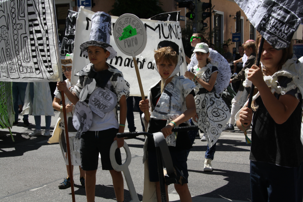 Das Foto zeigt kostümierte Kinder mit Schildern und Transparenten bei einer Parade.