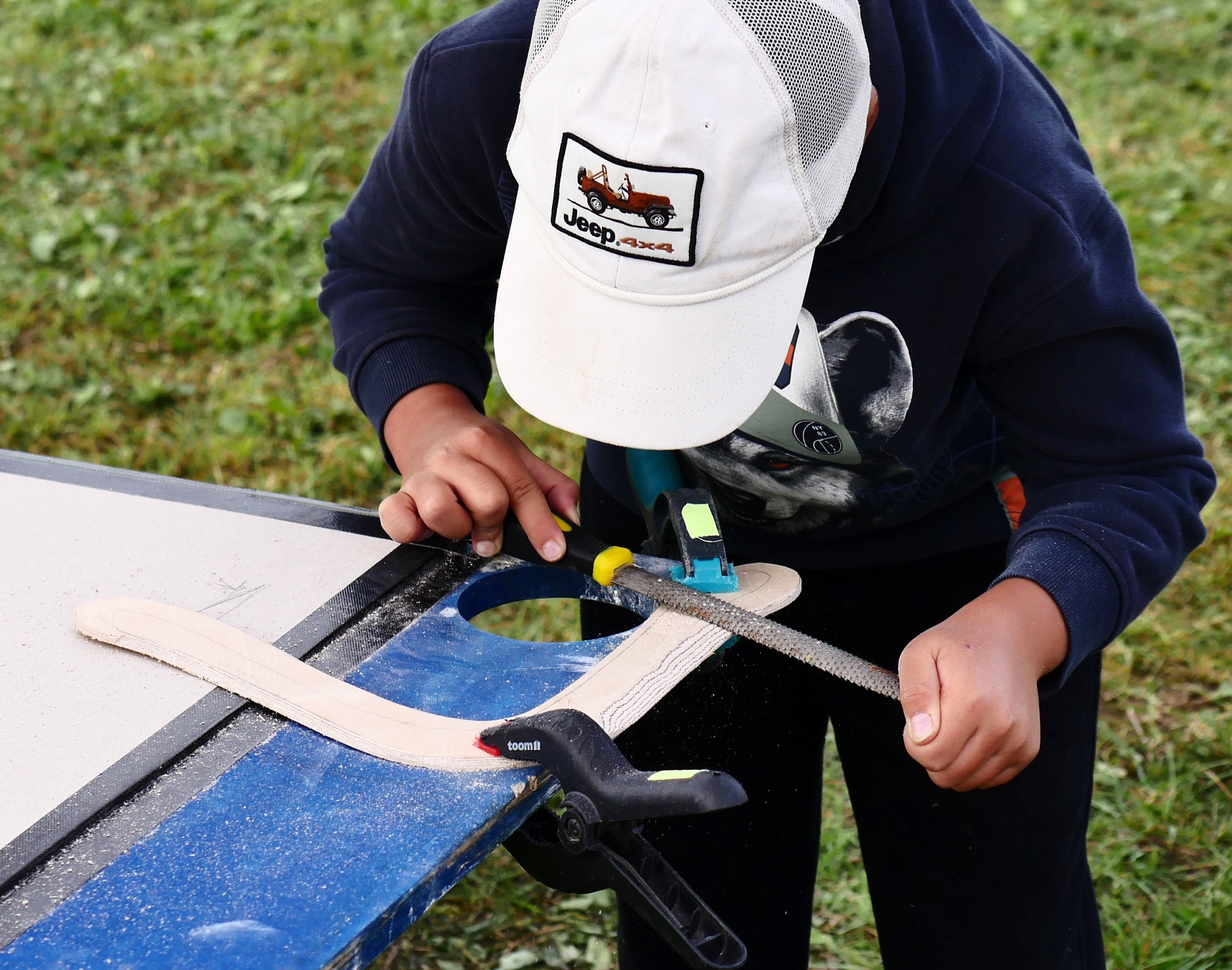 Drachenfest_Bumerangbau_c AKapfhammer Ein Kind arbeitet im Freien intensiv an einem Bumerang aus Holz.