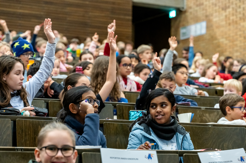 In einem Hörsaal sind Kinder zu sehen, von denen sich einige mit Handzeichen melden.