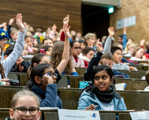 In einem Hörsaal sind Kinder zu sehen, von denen sich einige mit Handzeichen melden.